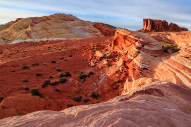 Arkasında kırmızı bir uçurum olan kayalık bir çöl manzarası. Güneş batıyor, sahne üzerine sıcak bir parıltı saçıyor.