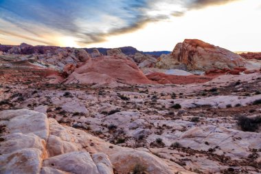 Arkasında güzel bir günbatımı olan kayalık bir çöl manzarası. Gökyüzü bulutlarla dolu, sakin ve huzurlu bir atmosfer yaratıyor. Kayalar tüm bölgeye dağılmış.