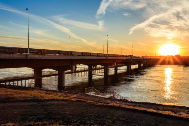 Arka planda günbatımı olan bir nehir üzerinde köprü. Köprü güneş tarafından aydınlatılır ve sıcak ve huzurlu bir atmosfer yaratır.