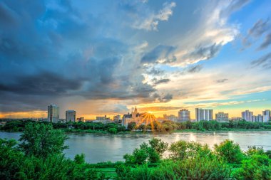 İçinden nehir akan bir şehir silueti. Gökyüzü bulutlu ve güneş batıyor.