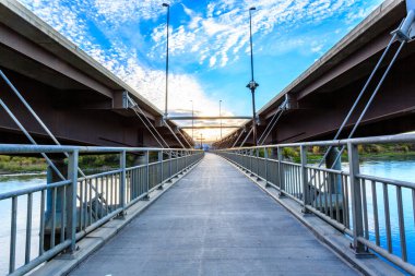 İki tarafında korkuluk olan bir köprü. Korkuluklar gümüş ve köprü uzun. Gökyüzü mavi ve bulutlar var