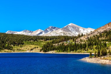 Önünde göl olan güzel bir dağ sırası. Gökyüzü açık ve mavi ve dağlar karla kaplı. Sahne huzurlu ve sakin, su dağları yansıtıyor.