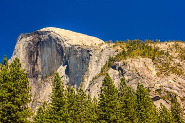 Tepesinde büyük bir kaya olan bir dağ ve etrafı ağaçlarla çevrili. Gökyüzü açık ve mavi