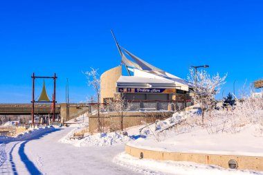 A winter scene with a snow-covered park and a building in the background. The building has a sign that reads 