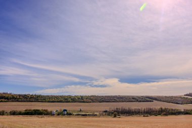 Uzakta bir ahır ve birkaç evi olan büyük bir tarla. Gökyüzü çoğunlukla açık ve birkaç bulut etrafa saçılmış.