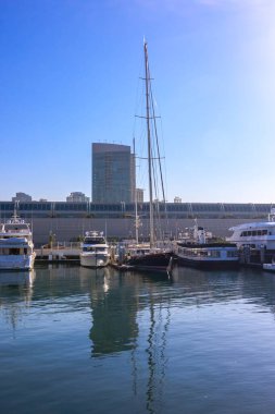 Bir grup tekne bir marinaya yanaşmış, biri de yelkenli. Su sakin ve gökyüzü berrak. Huzurlu ve sakin bir atmosfer yaratıyor.