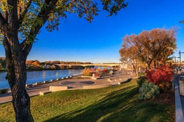 İçinden nehir akan bir park ve arka planda bir köprü. Gökyüzü mavi, ağaçlar turuncu ve kırmızı.