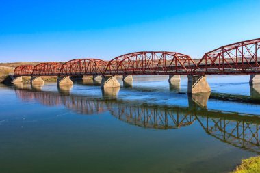 Kırmızı bir köprü, köprünün kırmızı rengini yansıtan suyla bir nehri kaplar. Köprü yeşil çimenli bir alanla çevrilidir.