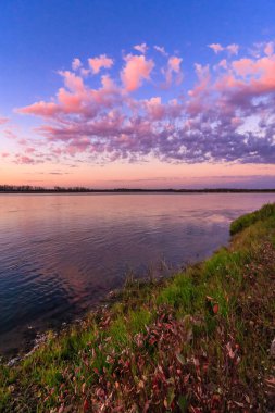 Gökte birkaç bulut olan bir gölün üzerinde güzel bir günbatımı. Gökyüzü pembe ve turuncu renklerin bir karışımıdır. Huzurlu ve huzurlu bir atmosfer yaratır. Su sakin ve durgun.
