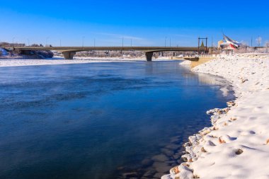 Köprü, diğer tarafında karlı bir kıyıya sahip bir nehri kapsıyor. Su donmuş ve gökyüzü berrak.