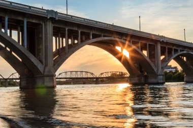 Köprü, arka planda günbatımı olan bir nehri kapsar. Köprü çok fazla detayla büyük bir yapıdır.