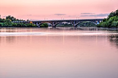 Bir köprü arka planda güzel bir günbatımı olan bir nehri kapsar. Köprü ışıklarla aydınlatılıyor, huzurlu ve huzurlu bir atmosfer yaratıyor.