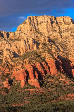 Arka planda bulutlu bir gökyüzü olan bir dağ sırası. Dağlar kırmızı kayalarla kaplıdır ve gökyüzü mavi ve beyaz karışımıdır.