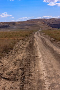 Çölün ortasında toprak bir yol esiyor. Yol asfaltsız ve üzerinde bir sürü kaya ve toprak var. Gökyüzü mavi ve arka planda bazı bulutlar var. Sahne sessiz ve huzurlu.