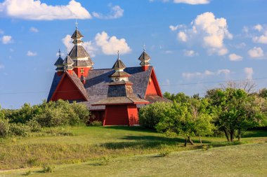 Beyaz çatılı kırmızı bir ev çimenli bir tepede oturur. Ev ağaçlar ve çalılarla çevrili, ona huzurlu ve huzurlu bir atmosfer sağlıyor. Gökyüzü açık ve mavi