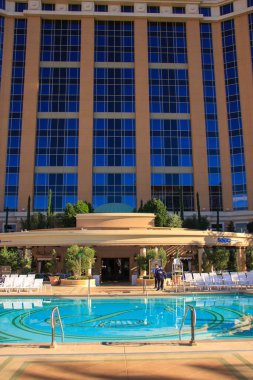 Arka planda mavi gökyüzü olan büyük bir otel havuzu. Havuz, şezlongları ve yemek masası olan bir veranda alanıyla çevrilidir.