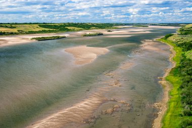 Kumlu bir kıyı şeridi ve arka planda birkaç ağaç olan bir nehir. Su sakin ve kıyı şeridi kayalarla kaplı.