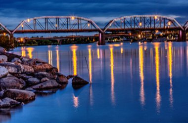 Bir köprü gece boyunca bir nehri kaplar, ışıklar suyu yansıtır. Sahne sakin ve huzurlu, köprü ve su bir huzur hissi yaratıyor.