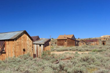Bir grup eski ahşap ev mavi gökyüzü olan bir tarlada. Evler eski ve terk edilmiş. Sahneye yalnızlık ve boşluk hissi veriyor.