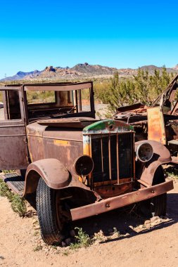 Çölde eski bir araba duruyor. Araba paslanmış ve yeşil süslemesi var. Çöl manzarası çorak ve ıssız.