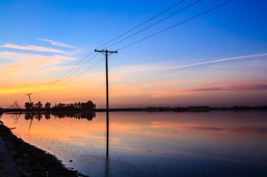 Bir elektrik hattı direği bir su kütlesinin önünde duruyor. Gökyüzü turuncu ve mavi renklerin güzel bir karışımıdır. Huzurlu ve huzurlu bir atmosfer yaratır.