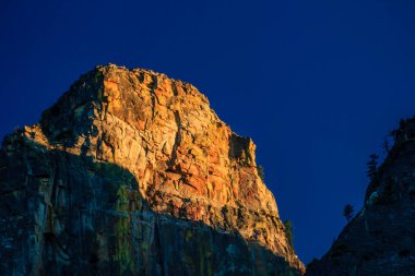 Kırmızı ve sarı kaya yüzeyli bir dağ güneş tarafından aydınlatılır. Gökyüzü mavi ve berrak, ve güneş uzaklardan batıyor.