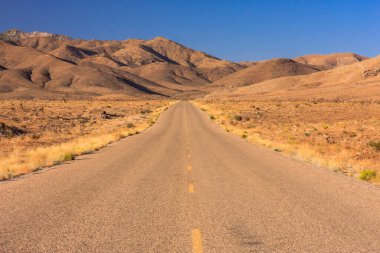 Arka planda dağlar olan çölde uzun bir yol. Yol boş ve gökyüzü temiz.