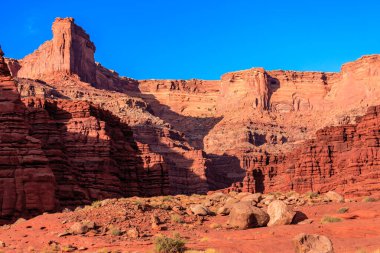 Arkasında büyük bir kaya oluşumu olan bir çöl manzarası. Gökyüzü açık ve mavidir, ve güneş parlıyor. Huzurlu ve huzurlu bir sahne.