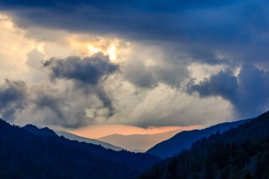 Gökyüzü bulutlu ve güneş batıyor. Dağlar arka planda ve bulutlar gökyüzünü kaplıyor.