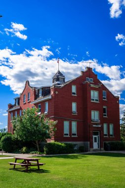 Üzerinde saat kulesi ve bayrağı olan kırmızı tuğlalı bir bina. Bina bir piknik masası ve bankı olan çimenli bir alanla çevrilidir. Gökyüzü açık ve mavi, birkaç bulut etrafa saçılmış