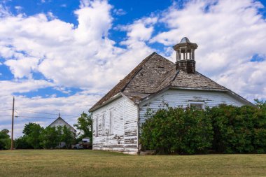 Çan kulesi olan küçük beyaz bir bina bir tarlada oturur. Bina ağaçlar ve çalılarla çevrili. Gökyüzü mavi ve arka planda bazı bulutlar var