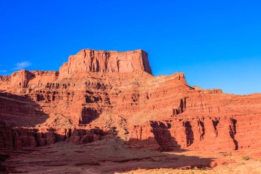 Üzerinde açık mavi gökyüzü olan büyük bir kırmızı kaya oluşumu çölde oturmaktadır. Güneş ışıl ışıl parlıyor, manzaraya sıcak bir parıltı saçıyor. Huzurlu ve huzurlu bir sahne.