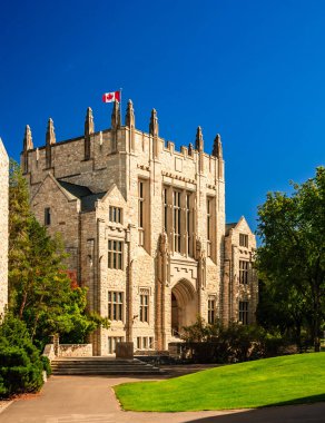 Üzerinde bayrak olan büyük bir taş bina. Bina ağaçlar ve otlarla çevrili. Gökyüzü açık ve mavi