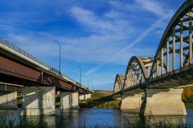Arka planda mavi gökyüzü olan bir nehrin üzerinde iki köprü. Soldaki köprü kırmızı çatılı beton bir köprü.