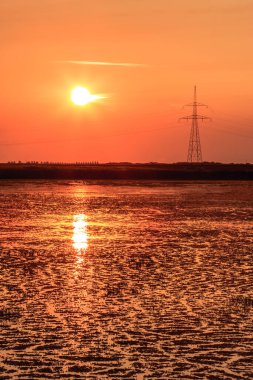 Arka planda elektrik hattı olan bir su kütlesinin üzerinde güzel bir günbatımı. Gökyüzü turuncu ve su sakin.