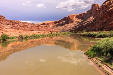 Arka planda bulutlu bir gökyüzü olan bir nehir bir kanyonun içinden akar. Su kahverengi ve bulanıktır ve gökyüzünün yansıması suda görülebilir. Huzurlu ve huzurlu bir sahne.