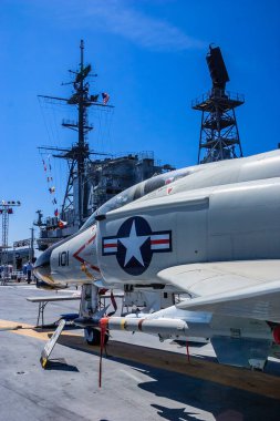 Bir savaş uçağı büyük bir geminin yanındaki pistte park halinde. Gökyüzü açık ve mavidir, ve güneş parlıyor. Güç ve güç kavramı