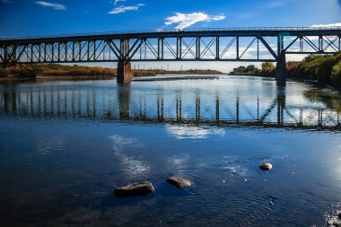 Bir köprü sakin mavi bir suyla bir nehri kaplar. Köprünün yansıması suda görülebilir.