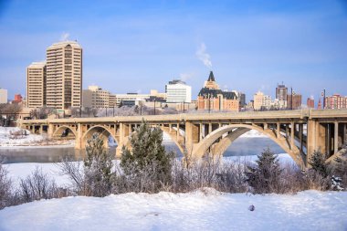 Arka planda bir şehir olan nehir üzerinde bir köprü. Gökyüzü açık ve güneş parlıyor.
