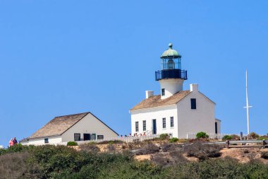 Arka planda mavi gökyüzü olan bir tepede bir deniz feneri var. Deniz feneri bir çitle çevrili ve birkaç kişi etrafında duruyor.