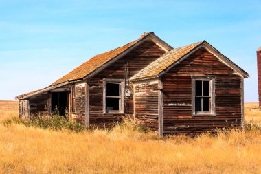 Çatı tarafı eğimli, köhne, eski bir ahşap ev uzun, kuru otlarla dolu bir alanda duruyor. Ev terk edilmiş ve bakımsız görünüyor, boyası soyulmuş ve camları kırılmış.