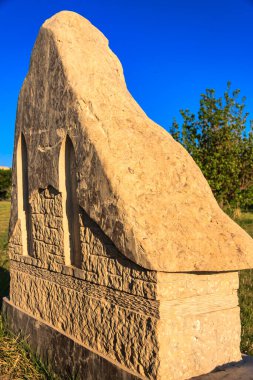 Üst tarafı eğimli büyük bir mezar taşı. Taş kahverengi ve yıpranmış görünüyor. Gökyüzü mavi ve berrak ve arka planda ağaçlar var.