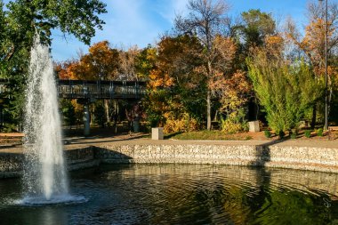 Bir çeşme gölete su püskürtüyor. Su yukarıdaki ağaçları ve gökyüzünü yansıtıyor. Huzurlu ve huzurlu bir sahne.
