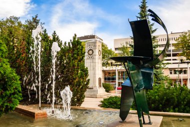 Parktaki bir çeşmenin önünde duran bir adam heykeli. Çeşme ağaçlarla çevrili ve üzerinde bir saat var. Heykel metalden yapılmıştır ve çeşmenin ortasına yerleştirilir.