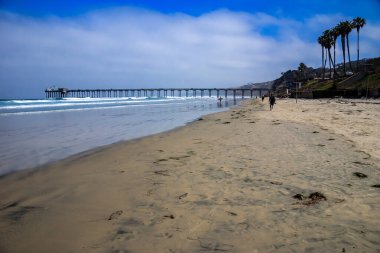 Arka planda palmiye ağaçları ve iskelesi olan bir sahil. Kıyı boyunca yürüyen birkaç kişi dışında sahil boş.