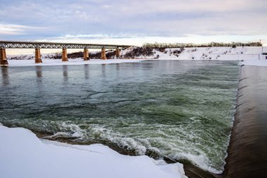 Bir köprü yeşilimsi bir renkle bir nehri gerer. Su dalgalı ve gökyüzü bulutlu.