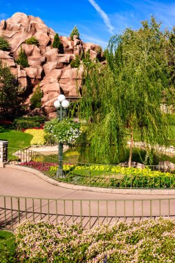 Taş duvarı ve yolu olan bir park. Yol ağaçlar ve çiçeklerle kaplı. Çiçekler sarı ve pembe.