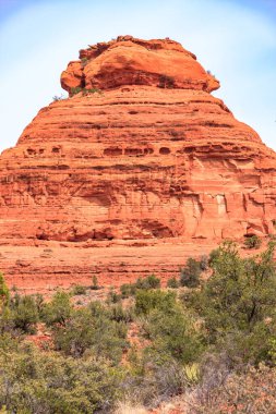 Önünde yeşil bir çalılık olan büyük bir kaya oluşumu. Kaya oluşumu turuncu ve gökyüzü mavi.