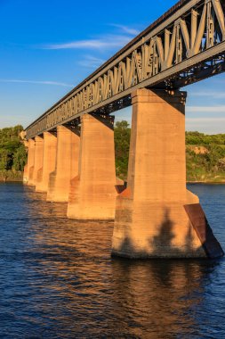 Arka planda mavi gökyüzü olan bir nehir boyunca uzanan bir köprü. Köprü beton ve çelikten yapılmıştır.