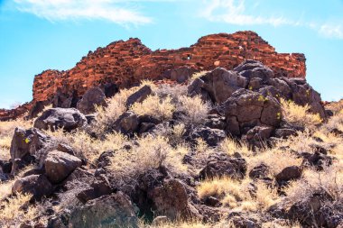 Uzakta büyük bir binası olan kayalık bir yamaç. Bina kırmızı tuğlalardan yapılmıştır ve birçok kayayla çevrilidir. Gökyüzü mavi ve arka planda bazı bulutlar var
